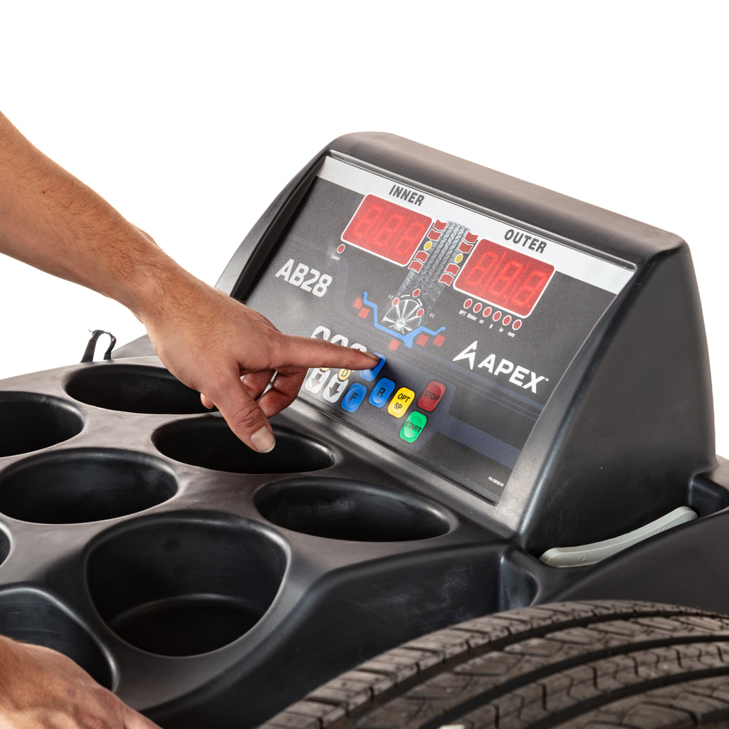 Technician operating the digital display and control panel above the integrated tool tray on an APEX AB28 wheel balancer.