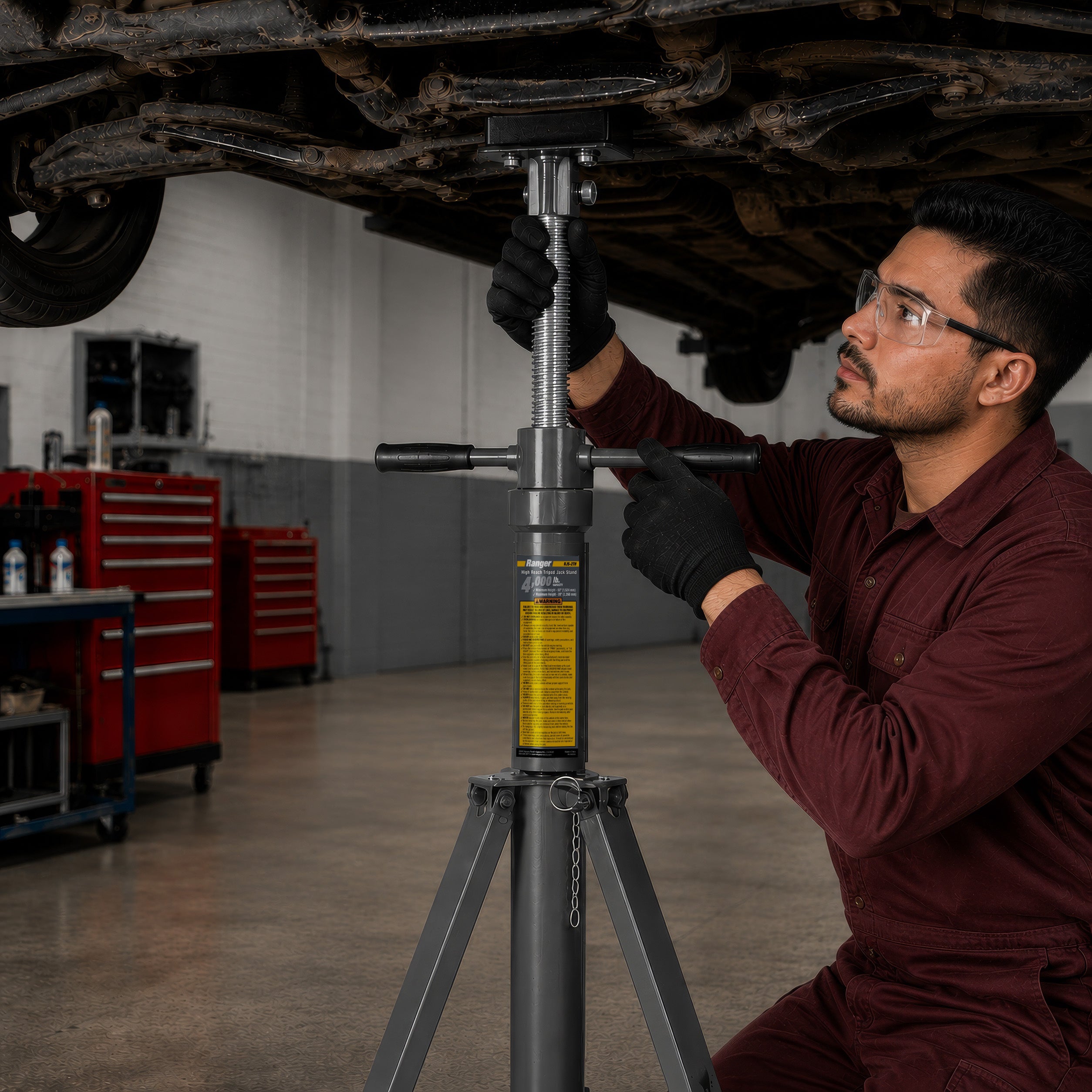 Technician working beneath a raised car supported by a Ranger RJS-2TH high-reach jack stand in a commercial automotive shop.